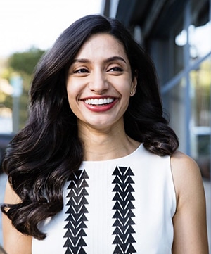 Woman smiling at camera wearing white sleeveless top with geometric pattern, outdoor street setting.