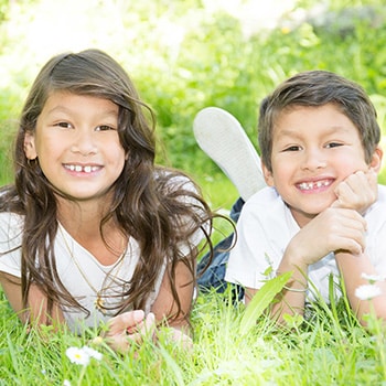 Two smiling children lying on grass outdoors with baseball bat and glove.
